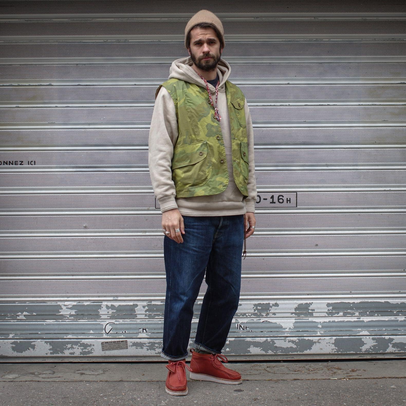 homme en sweat à capuche oversize avec un gilet de chasse camouflage militaire un bonnet ne laine et des clarks wallabee orange