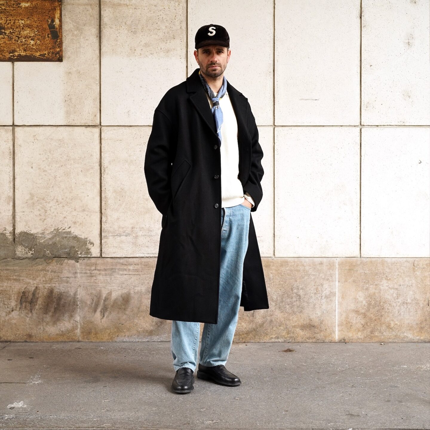 tenue pour l'hiver d'un homme avec un manteau long en laine noir, coupe oversize, porté avec une casquette noire supreme, un pull en lane mérinos Nitto, un jean délavé taile haute botali et des mocassins noirs